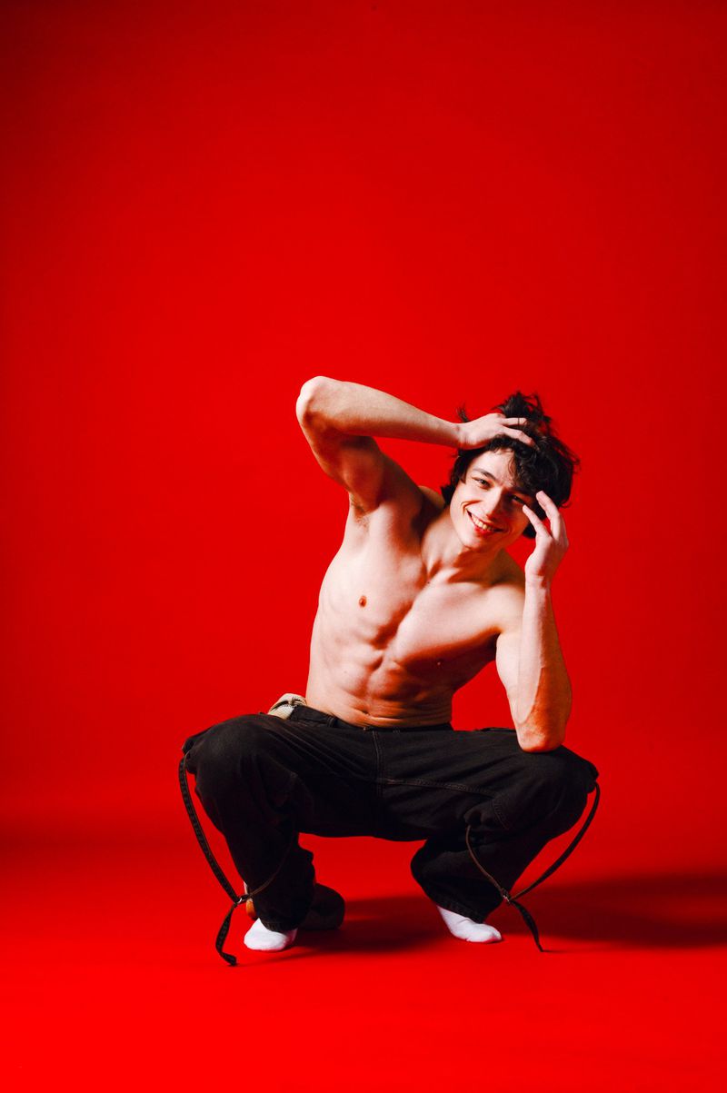 Athletic man with bare torso squats on a red background with his hands raised to his head and a happy smile.