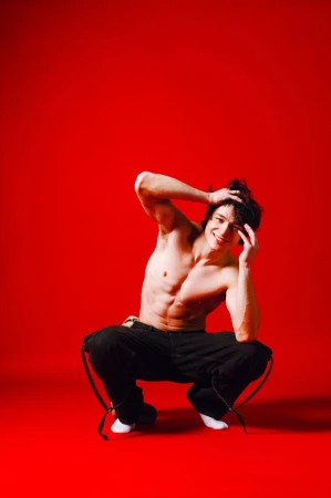 Athletic man with bare torso squats on a red background with his hands raised to his head and a happy smile.