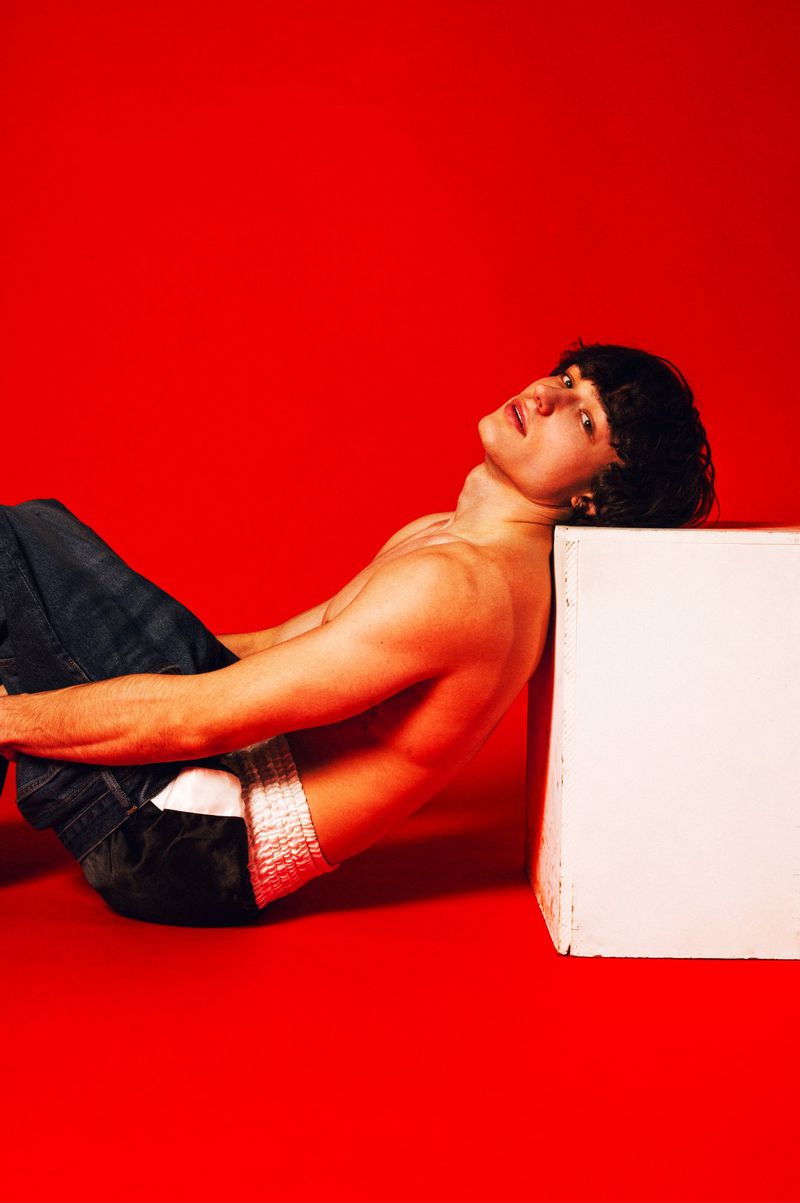 A shirtless young man in jeans leans back against a white cube, posing in a photo studio against a vivid red backdrop.
