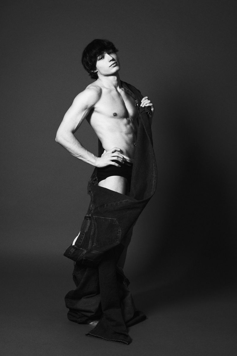 Monochrome studio shot of a slender adult male with dark hair, wearing denim overalls and briefs, posing with hand on hip, looking up.