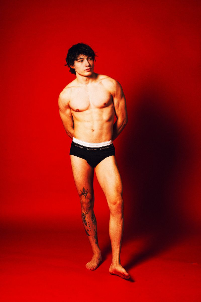 Athletic man poses in black briefs against a vibrant red background. Toned physique, dark hair, in a photo studio setting.