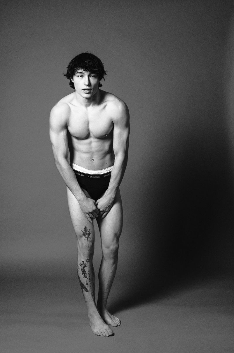 Black and white studio shot of a shirtless young adult male wearing briefs, standing barefoot with a slight bend at the knees.