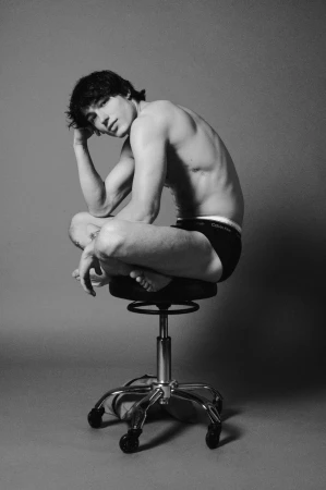 Monochrome studio portrait of a seated shirtless man posing on a rolling stool against a grey background.