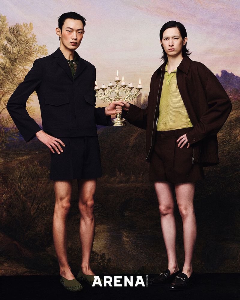 Two fashion models in dark blazers and shorts stand holding a candelabra against a backdrop resembling a landscape painting, indoors.