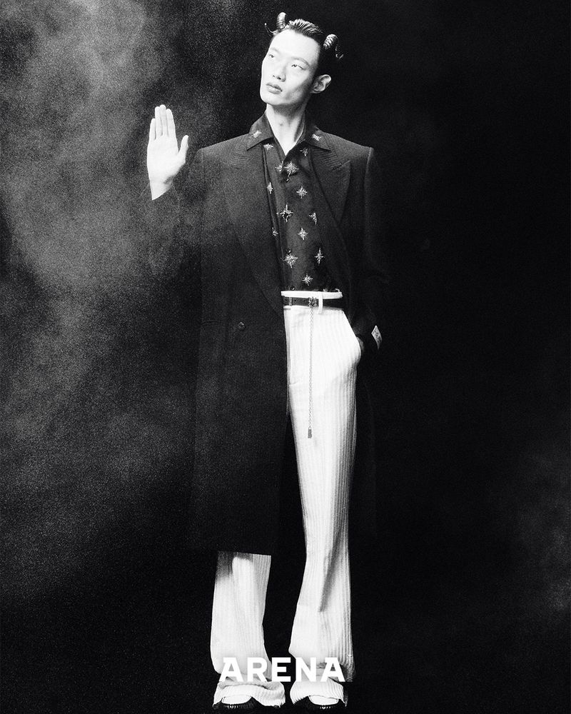 Monochrome fashion shot of an Asian male model in a long black coat and vertically striped pants against a dark, smoky background.