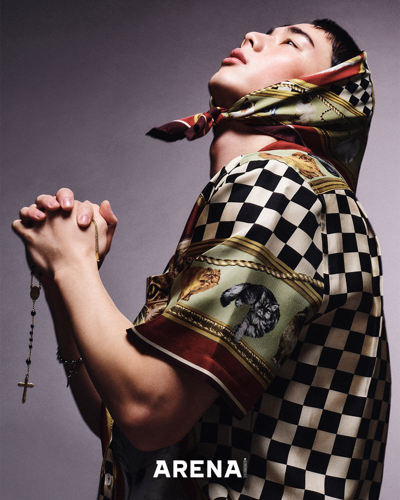 Portrait of an adult male fashion model with a patterned headscarf, hands clasped, praying, wearing a patterned shirt and necklace.