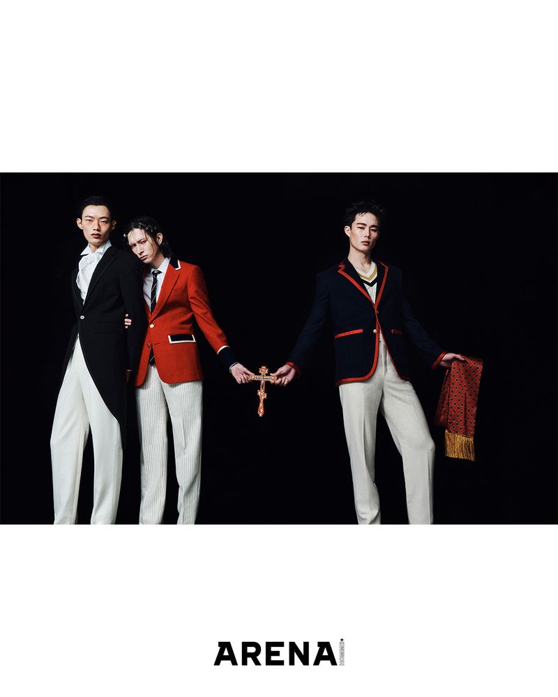 Three Asian male fashion models wearing formal jackets and white pants, holding a gold crucifix against a dark studio backdrop.