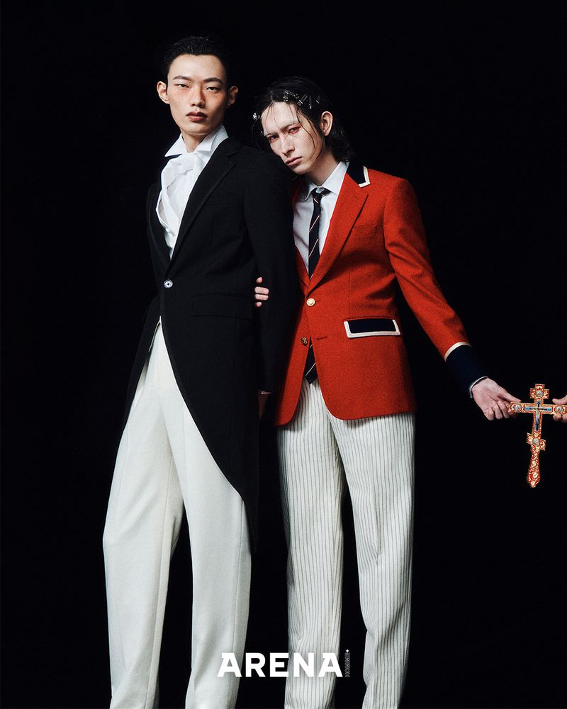 Two men pose in formal wear against a black backdrop, one in a black tailcoat, the other in a red jacket, for a fashion editorial.
