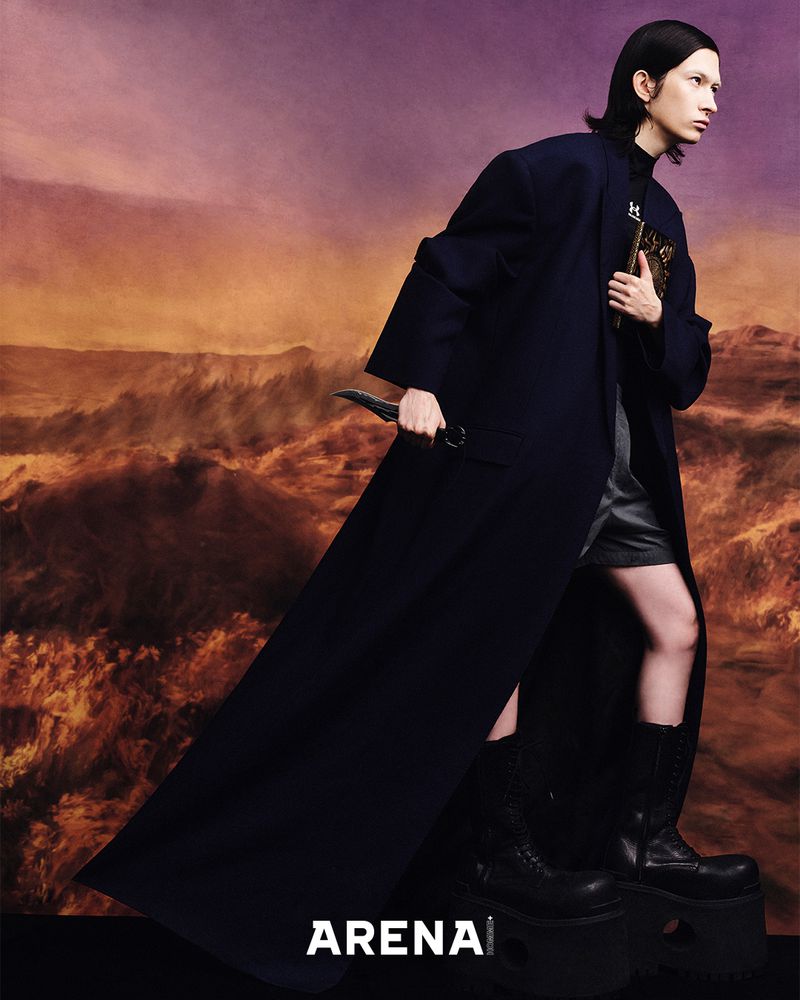 Fashion shot of a male model wearing a long dark coat, shorts, and platform boots while holding a knife, set against a desert backdrop.