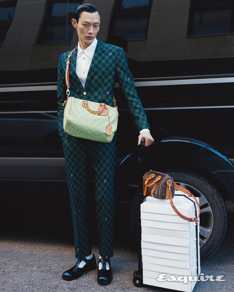 Asian man in patterned suit stands with luggage by a black vehicle, carrying branded bag, stylish street fashion editorial.