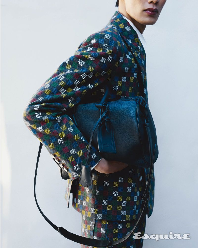 A man in a colorful patterned suit holds a black leather handbag against a clean backdrop, showcasing fashion and style.