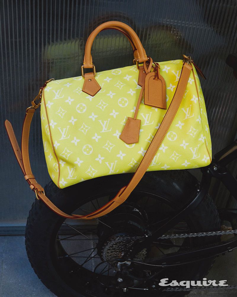 A yellow Louis Vuitton Speedy bag with tan leather straps rests on a black bicycle tire against a neutral-toned backdrop.