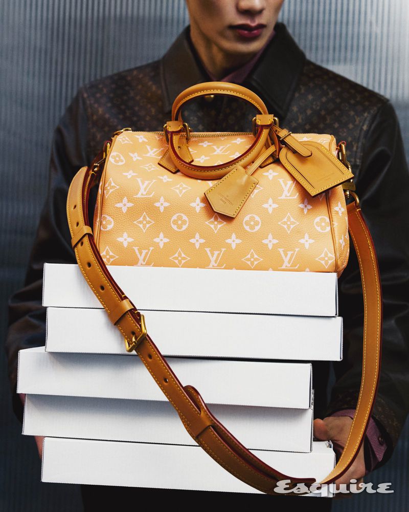 A person holds a light orange Louis Vuitton handbag with white logos, balanced on a stack of white boxes, against a textured wall.