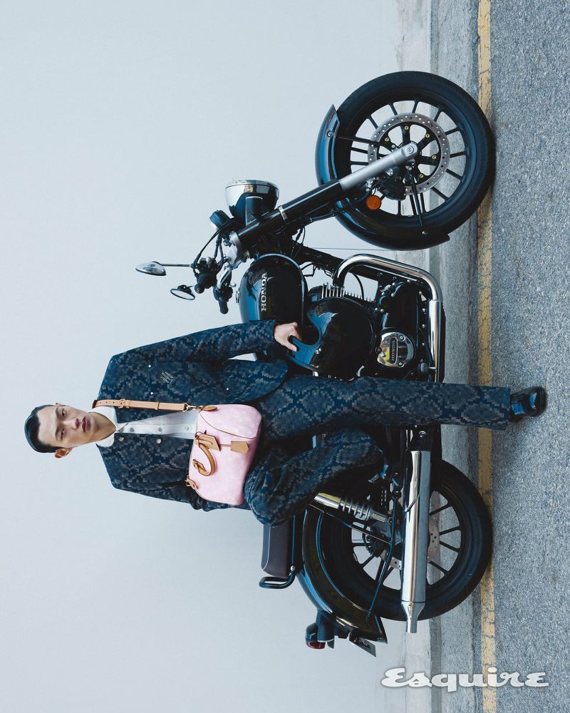 Stylish man in patterned suit poses on black motorcycle outdoors, holding a pink purse, editorial fashion shoot, neutral backdrop.
