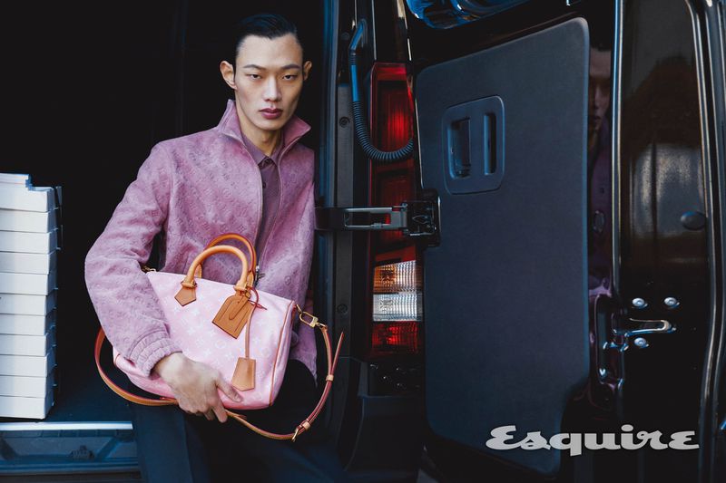 Asian male model poses with pink handbag at van door; wearing a patterned mauve jacket and standing in front of a black vehicle.