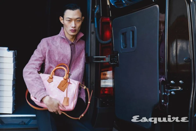 Asian male model poses with pink handbag at van door; wearing a patterned mauve jacket and standing in front of a black vehicle.