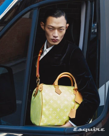 Asian man in a black velvet jacket, inside a car, showcasing a Louis Vuitton yellow monogrammed Speedy bag with a shoulder strap.