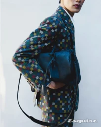 Model wearing a colourful patterned suit holding a dark leather duffel bag against a white background