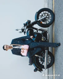 Model Hoyong Kim wearing a patterned suit and carrying a pink handbag, leaning on a dark motorcycle outdoors.