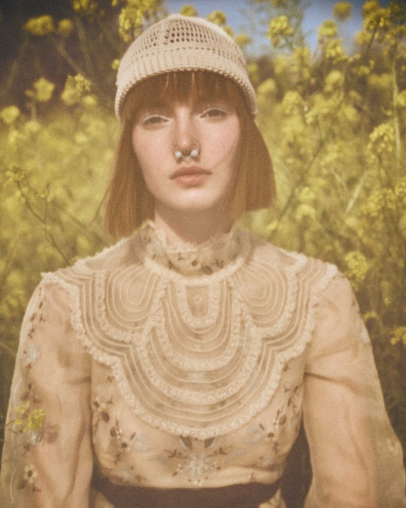 Close-up portrait of a woman in a lace top and knit cap, standing in a field of yellow flowers, with a shallow depth of field.
