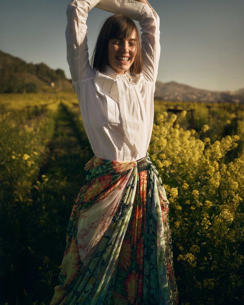 A cheerful woman in a white shirt and colorful floral skirt raises her arms in a sunlit field of yellow flowers with a blurred mountain backdrop.