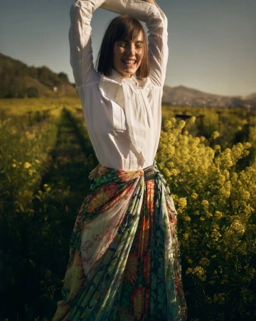 A cheerful woman in a white shirt and colorful floral skirt raises her arms in a sunlit field of yellow flowers with a blurred mountain backdrop.
