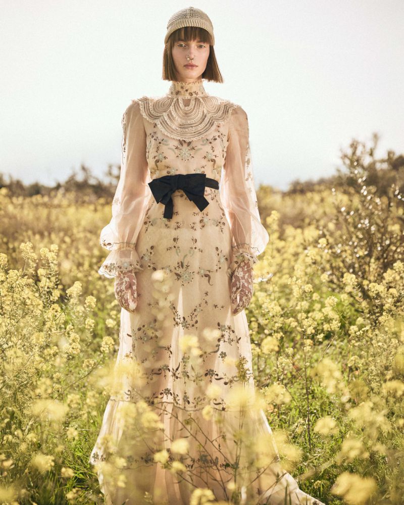 Fashion shot of a female model in a sheer floral dress and knitted cap standing in a field of yellow flowers against a soft sky.