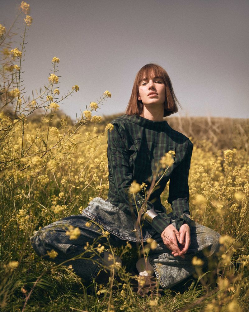 Woman in a plaid top crouches in a field of yellow flowers, creating a natural, warm scene with soft light and a serene atmosphere.