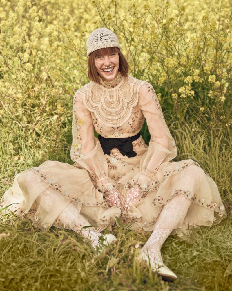 An adult female model sits in a field of yellow wildflowers, wearing a couture gown, knitted hat, and lace stockings, smiling at the camera.