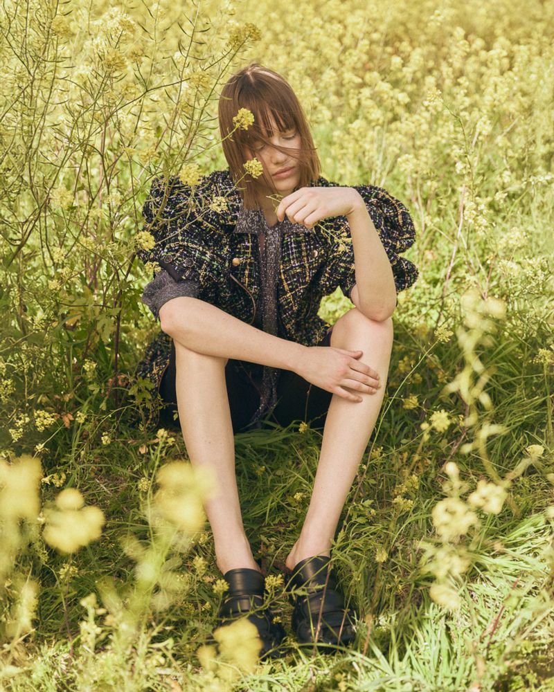 Woman in a textured jacket sits in a field of yellow wildflowers. She wears black loafers and a contemplative expression in natural light.