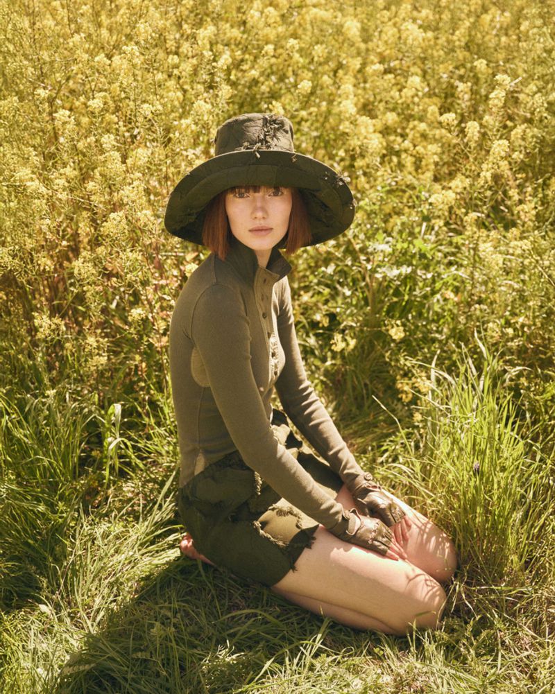 A woman in a green outfit and hat kneels in a sunlit field of yellow flowers and tall grass, creating a natural, outdoor fashion scene.