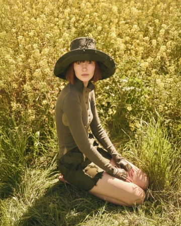 Model in olive green top and matching wide-brim hat kneeling in a lush meadow of wildflowers