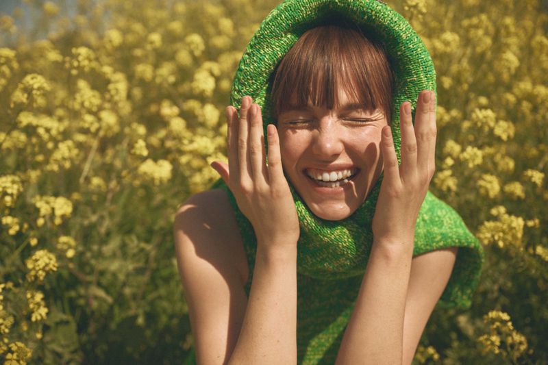 Close-up shot of a smiling woman with brown hair wearing a green hooded shawl, hands near her face, standing in a field of yellow flowers.