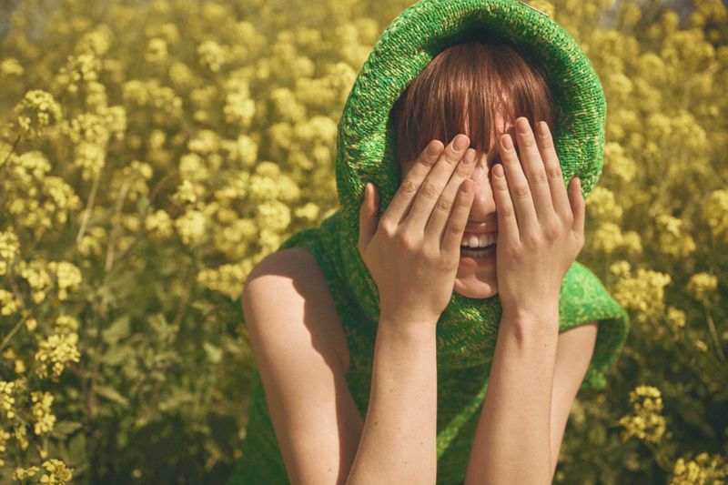 Woman with auburn hair covers face with hands in outdoor field of yellow wildflowers, wearing a green hooded top and smiling broadly.
