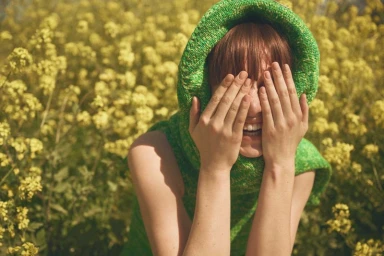 Model wearing a bright green knitted hood covering her head while hiding her face with both hands amid yellow wildflowers.