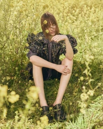 Model seated amidst tall wild yellow mustard flowers wearing a patterned tweed jacket with puffed sleeves.