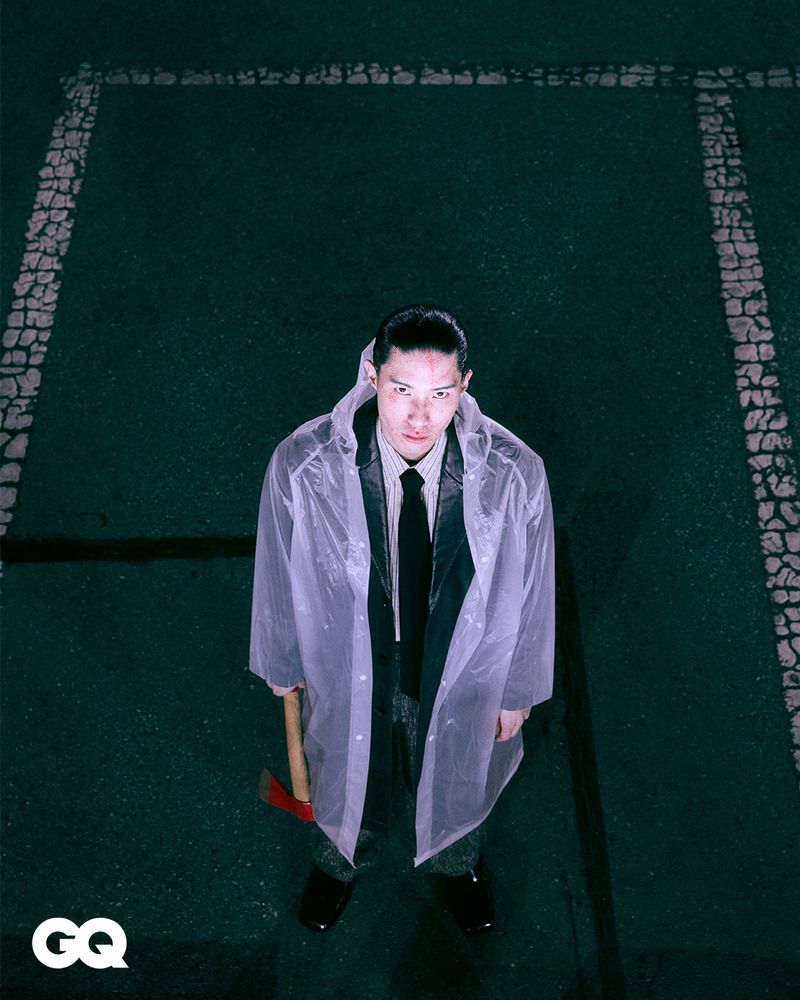 Adult man in a translucent raincoat and holding an axe stands in an outdoor setting, looking down at the camera with a serious expression.