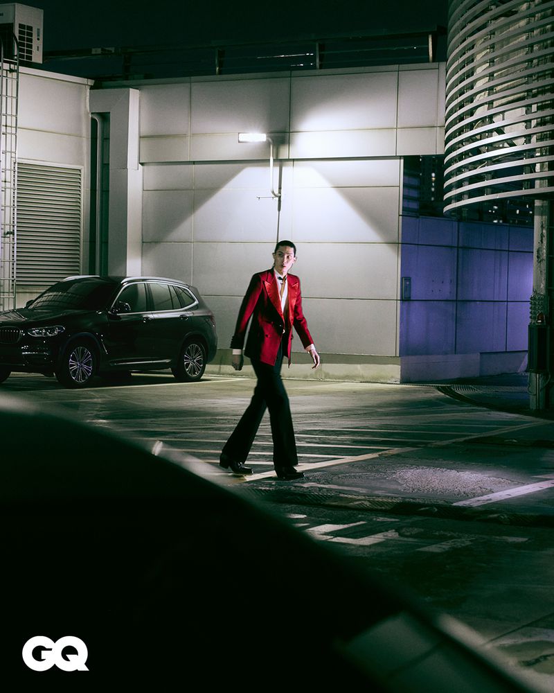 Man in a red blazer walks across a crosswalk at night with a black SUV and a modern building visible in the urban background.