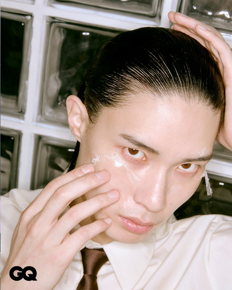Close-up portrait of a young adult male applying facial cream. He has dark hair and is wearing a collared shirt and tie.