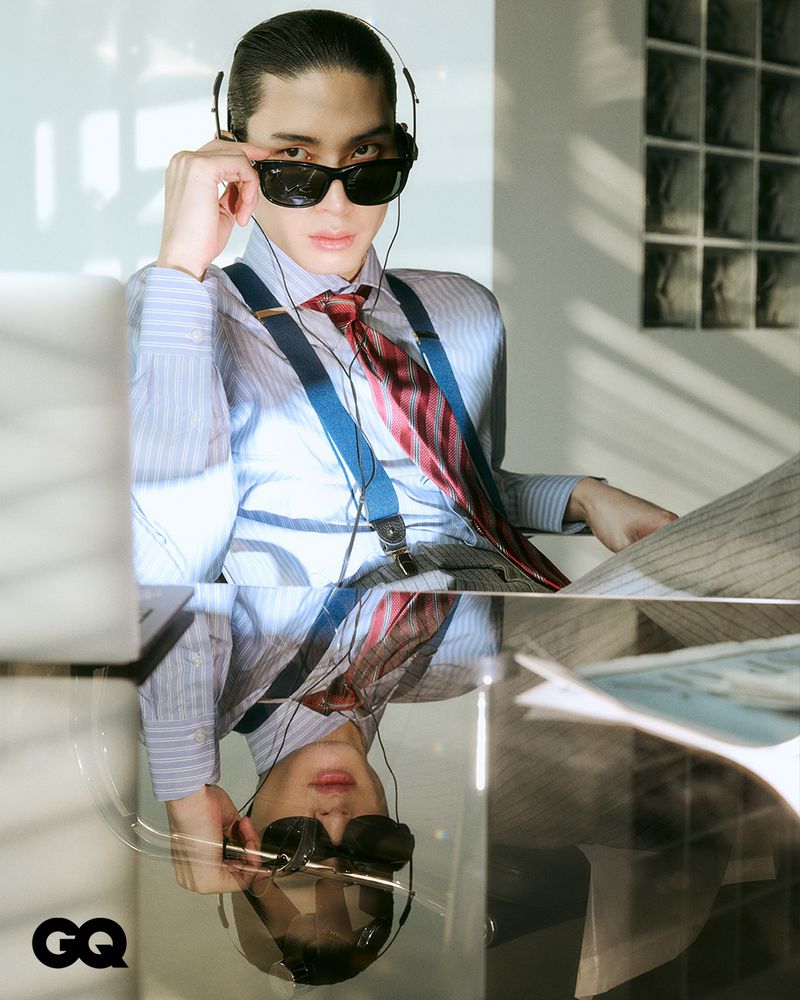 Stylish adult male poses indoors wearing sunglasses, headphones, pinstripe suit, suspenders, and a patterned tie while seated at a glass table.