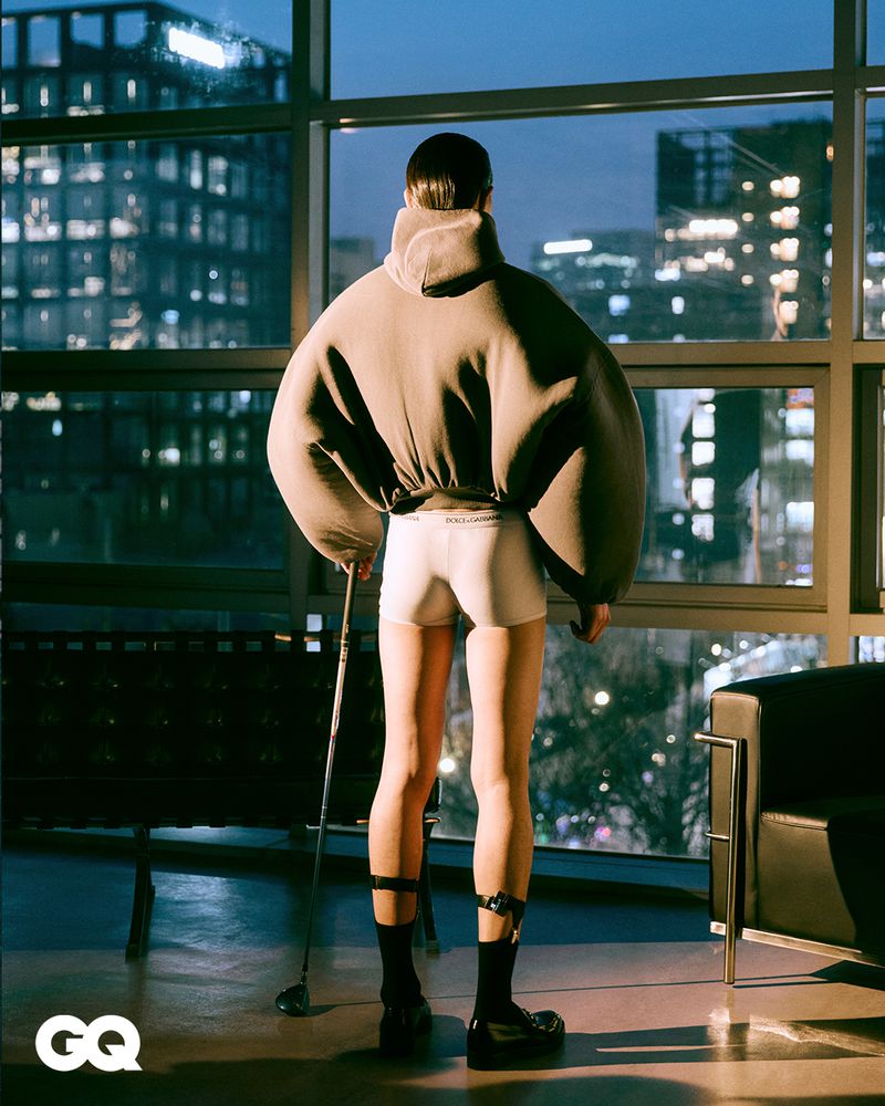 Back view of a person with a golf club in an oversized hoodie and briefs, set against a high-rise city building backdrop at twilight.