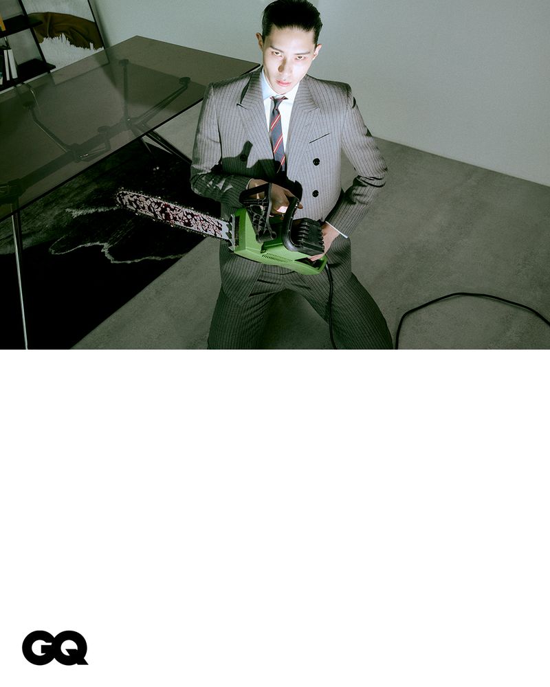 Adult male in a grey pinstripe suit sits holding a green chainsaw while posing in front of a white wall next to a modern table.