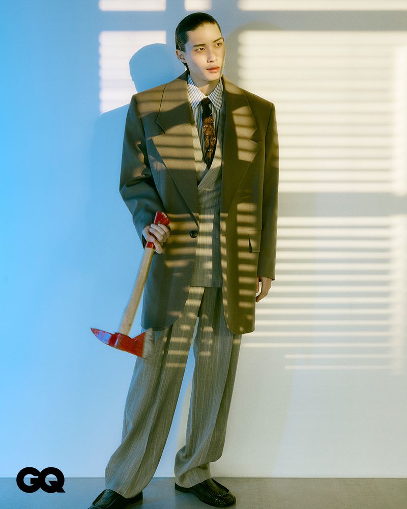 An adult male in an oversized olive suit stands indoors with an axe, illuminated by window-blind light, in a fashion editorial with GQ logo.