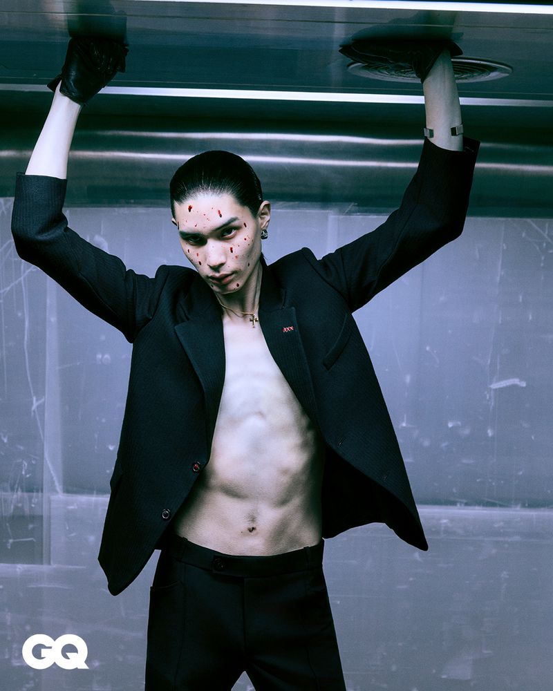 Close-up of an adult male fashion model in a black suit, bare torso, theatrical makeup, and black gloves posing indoors with a metal background.