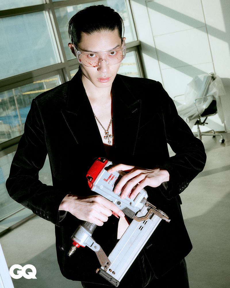 Portrait of a young adult male in a black suit and safety glasses, holding a nail gun, standing indoors with a window in the background.