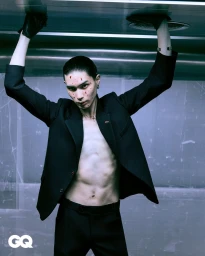 Model with slicked-back hair and markings on face wearing an open black blazer and trousers, holding up arms against a metallic ceiling.