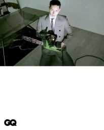 Man wearing a striped grey suit holding a green chainsaw inside a modern room with a glass table.