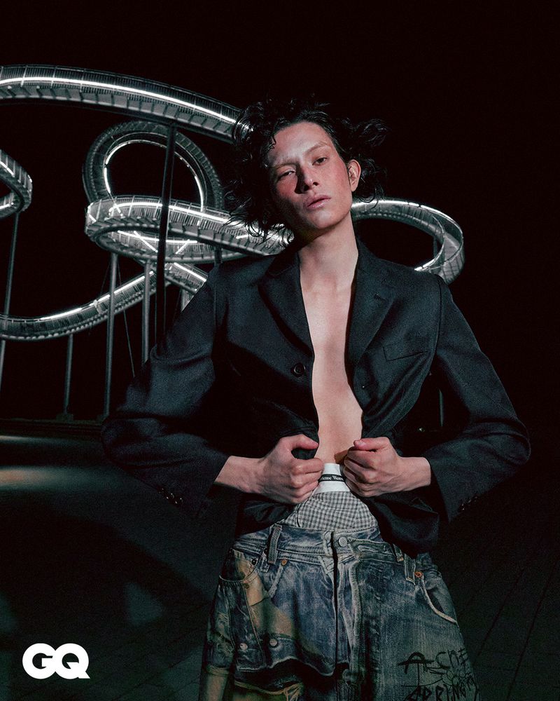 Editorial fashion shot of a slender adult man wearing a dark blazer and distressed denim, standing in front of a coiled metal structure.