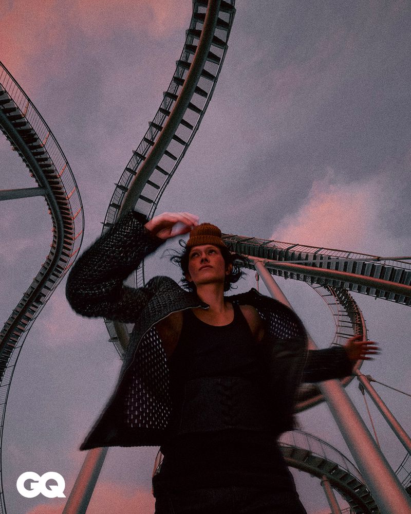 A young adult man in stylish jacket and beanie strikes a pose in front of a winding roller coaster against a dramatic cloudy sky.