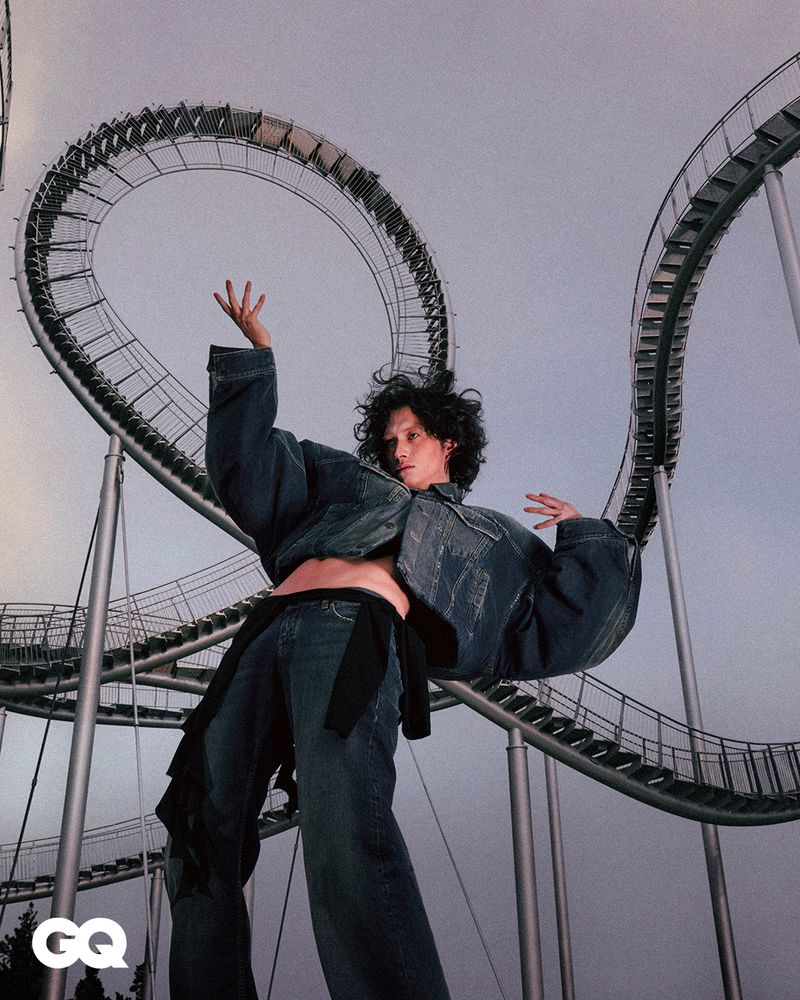 A young adult poses on a roller coaster wearing an oversized denim jacket and matching jeans with GQ logo bottom left.